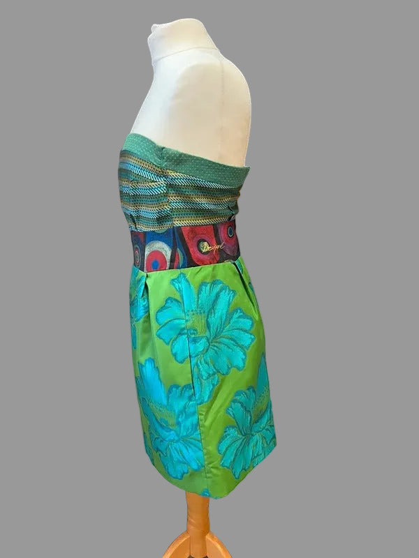 A colorful strapless dress displayed on a white dress form mannequin. The dress features a green and blue floral patterned skirt and a striped bodice with a multicolor patterned waistband, set against a white wall and black metal shutter background.