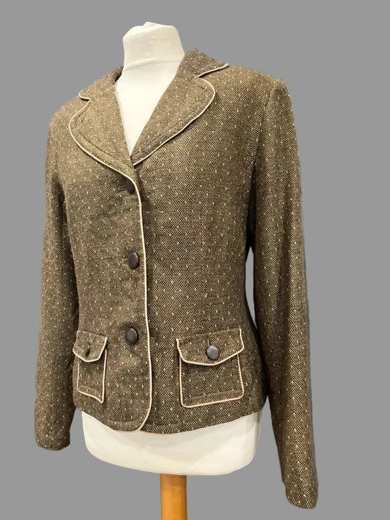 A brown tweed blazer jacket with beige contrast piping and dark buttons, displayed on a white mannequin torso against a gray paneled wall.