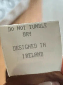 A close-up of a person's hand holding a brown garment, showing a sewn-on paper label that reads 'DO NOT TUMBLE DRY DESIGNED IN IRELAND'.