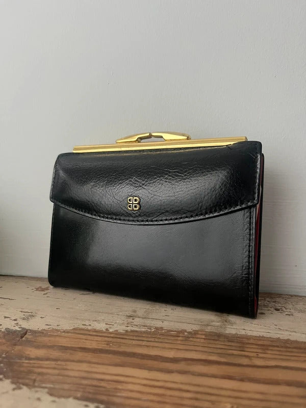 A black leather clutch wallet with a gold-tone metal frame and a gold logo emblem on the front, resting on a rustic wooden surface against a plain light gray wall.