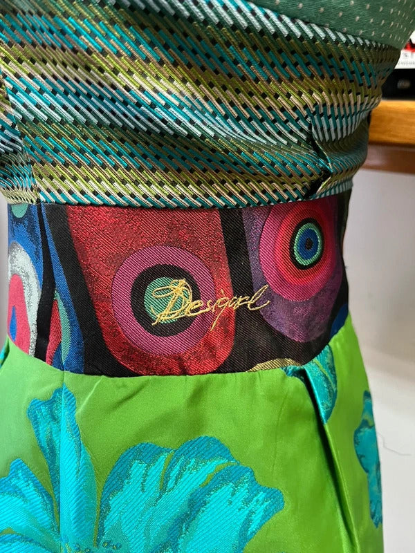Close-up of a colorful garment featuring a patterned waistband with circular motifs and a bright green lower section with blue floral embroidery, branded with 'Desigual'