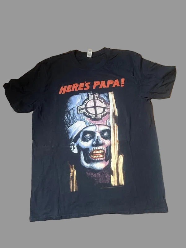 A black short-sleeve graphic t-shirt laid flat on a wooden floor. The shirt features the text 'HERES PAPA!' in red at the top, above a graphic of a zombie skull wearing a bandana with a crosshair symbol.
