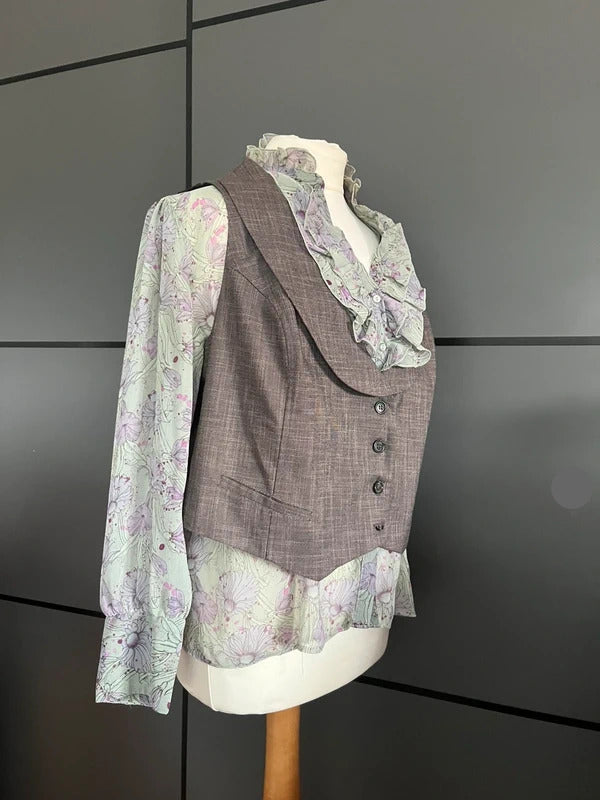 A women's vintage-style outfit displayed on a mannequin, featuring a taupe-brown vest over a long-sleeve ruffled blouse with a light purple and green floral print.