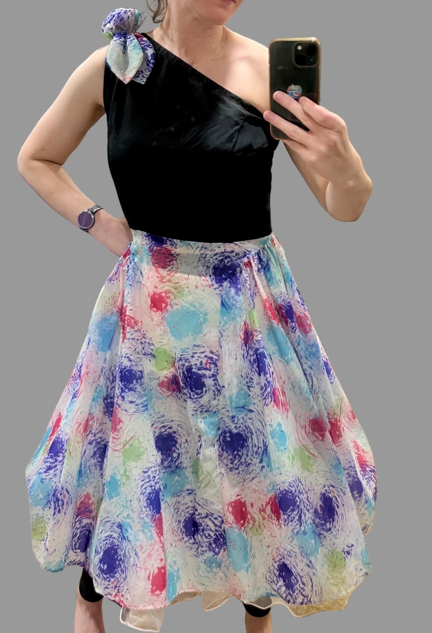 Woman taking a mirror selfie wearing a black top and colorful skirt.